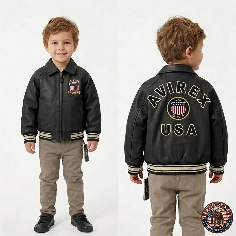 Puede incluir: Chaqueta de cuero negra con el texto "AVIREX USA" en la espalda y un parche en la parte delantera. La chaqueta tiene cuello, cremallera y pu&ntilde;os y dobladillo a rayas. El ni&ntilde;o lleva pantalones color caqui y zapatos negros.