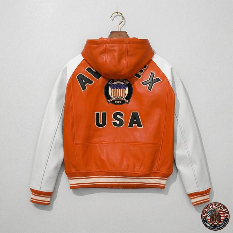 K&ouml;nnte beinhalten: Orange-wei&szlig;e Lederjacke mit Kapuze. Auf dem R&uuml;cken steht das Wort "USA" in Schwarz, ein Aufn&auml;her mit "ALL AMERICAN" und "1975". Wei&szlig;e &Auml;rmel und ein gestreifter Saum. Klassischer Varsity-Stil.