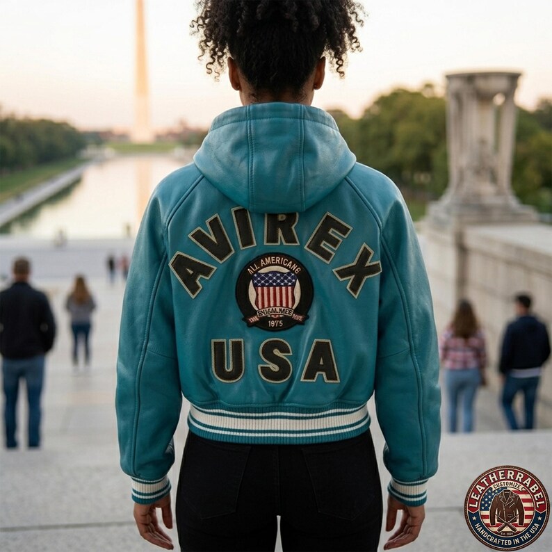 K&ouml;nnte beinhalten: Eine t&uuml;rkisfarbene Lederjacke mit Kapuze und dem Schriftzug AVIREX USA in gro&szlig;en dunklen Buchstaben auf dem R&uuml;cken. Ein runder Aufn&auml;her in der Mitte zeigt eine US-Flagge mit dem Text ALL AMERICANS 1975. Der Saum ist gerippt und gestreift.