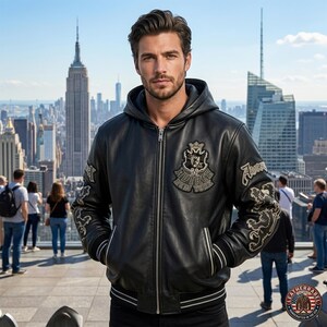 May include: A black leather hooded jacket featuring a front zipper closure, ribbed cuffs with white stripes, and embroidered patches on the chest and sleeves. The chest patch displays a crown and the text Avirex New York in a crest design.