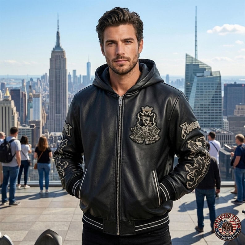 May include: A black leather hooded jacket featuring a front zipper closure, ribbed cuffs with white stripes, and embroidered patches on the chest and sleeves. The chest patch displays a crown and the text Avirex New York in a crest design.
