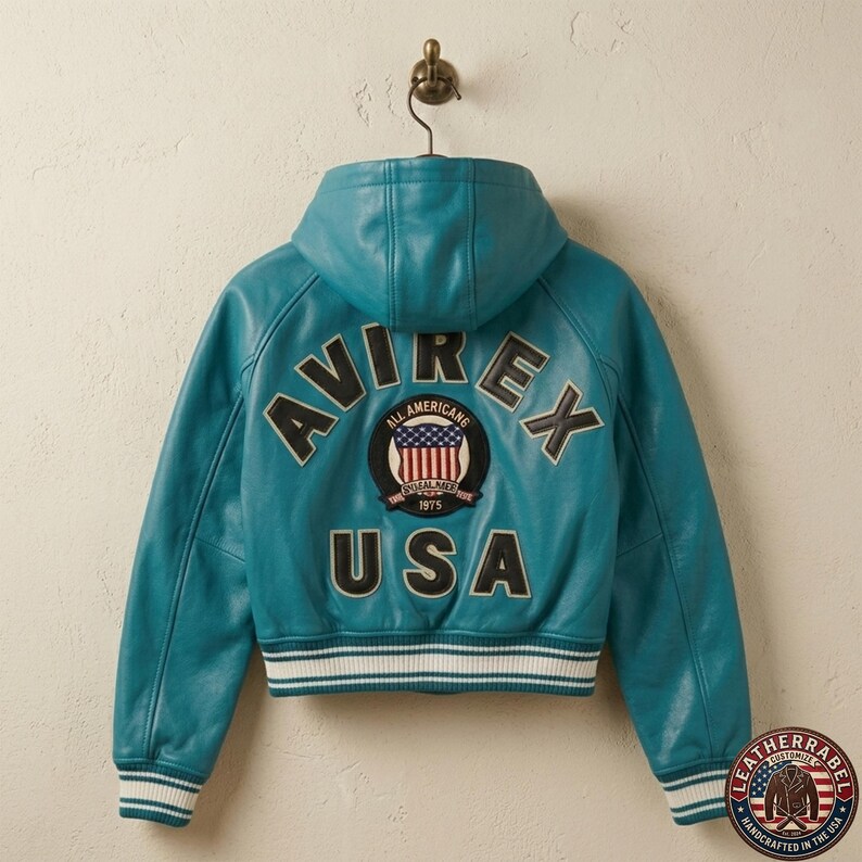 K&ouml;nnte beinhalten: Eine t&uuml;rkisfarbene Lederjacke mit Kapuze und dem Schriftzug AVIREX sowie USA in schwarzen Buchstaben auf der R&uuml;ckseite. Ein runder Aufn&auml;her in der Mitte zeigt eine amerikanische Flagge mit dem Text ALL AMERICANS 1975. Gerippte B&uuml;ndchen und Saum.