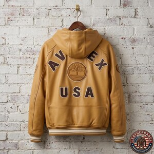 May include: A tan leather jacket with a hood, hanging on a wooden hanger against a white brick wall. The back of the jacket features the word "AVEX" above "USA", with a tree logo. The jacket has a ribbed hem and cuffs.
