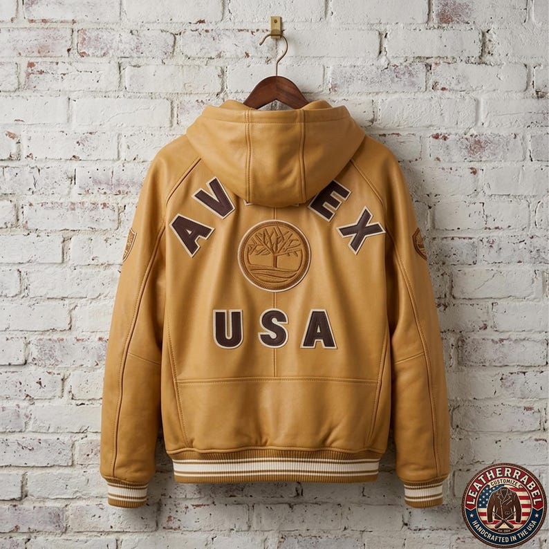 May include: A tan leather jacket with a hood, hanging on a wooden hanger against a white brick wall. The back of the jacket features the word "AVEX" above "USA", with a tree logo. The jacket has a ribbed hem and cuffs.