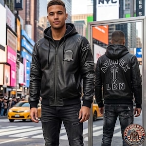 May include: Black leather jacket with a hood, zipper closure, and embroidered details. The jacket features the "AVIREX" logo on the chest and back, with "LDN" below. The model is wearing black jeans in a city setting.