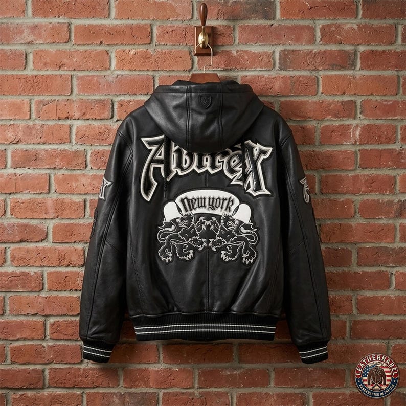 May include: A black leather hooded jacket featuring white embroidered lettering that reads Aubrey and New York, accompanied by a graphic of two lions. The jacket includes ribbed cuffs and a hem with white stripes, showcasing detailed stitching.