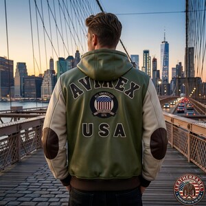 May include: An olive green hooded leather jacket with cream-colored sleeves and dark brown elbow patches. The back features the text AVIREX in an arch, a circular emblem with a US flag, and the text USA. The jacket has a ribbed hem and cuffs.