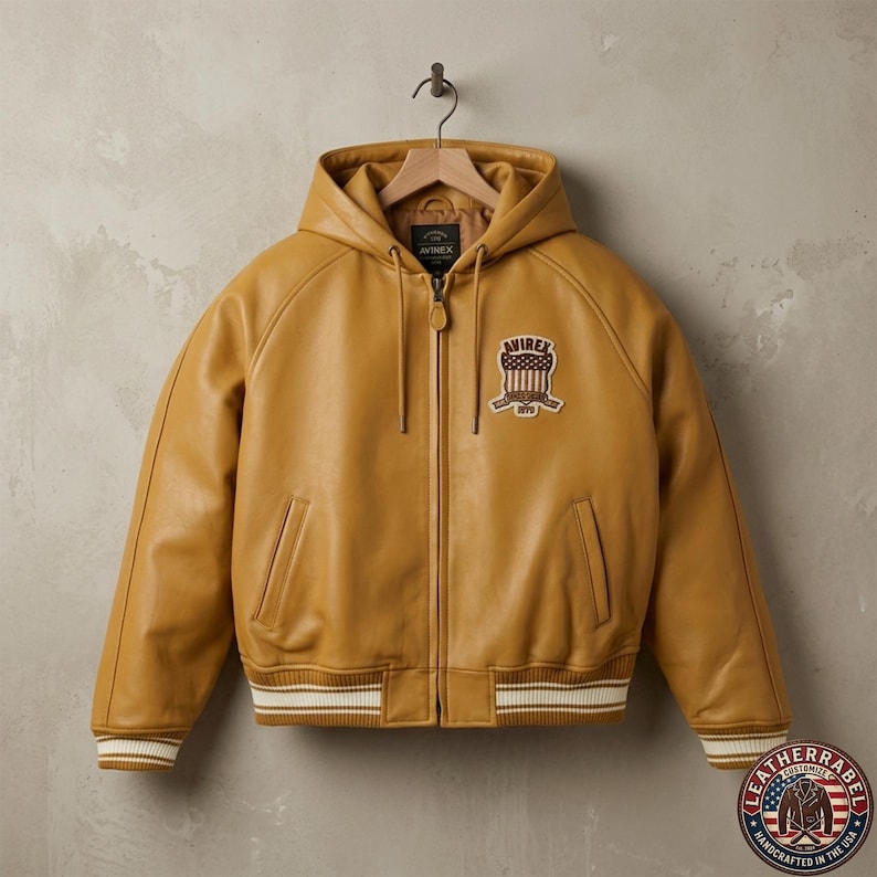 May include: Mustard yellow leather jacket with a hood, zipper closure, and ribbed cuffs and hem with white stripes. Features a patch with the text "AVIREX" and a logo. The jacket is hanging on a wooden hanger.