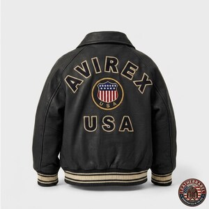 Puede incluir: Chaqueta de cuero negra con la palabra "AVIREX" arqueada sobre los hombros y "USA" debajo de un emblema circular. El emblema presenta un escudo con rayas rojas y blancas y estrellas azules. La chaqueta tiene cuello y pu&ntilde;os a rayas.