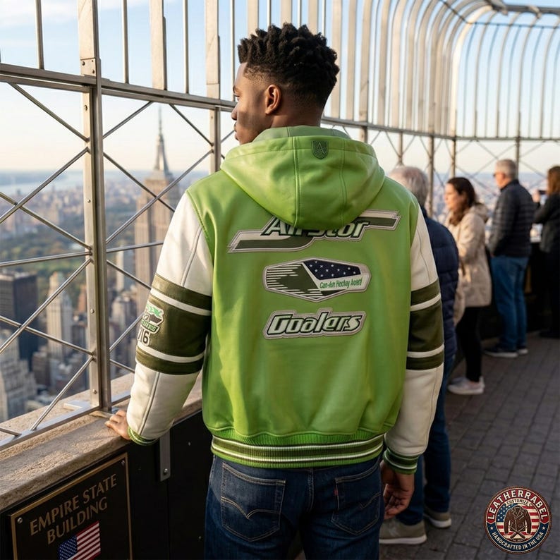 May include: A green and white leather jacket with a hood, featuring the text "Arcor Coolers" on the back. The jacket has white sleeves with green stripes and a patch on the left arm. The person is standing at the Empire State Building.