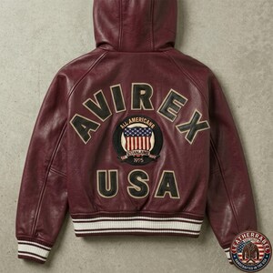 Puede incluir: Una chaqueta de cuero con capucha en color burdeos que presenta el texto AVIREX USA en letras grandes de color negro y crema en la espalda. Un parche circular en el centro muestra un gr&aacute;fico de la bandera estadounidense con el texto ALL AMERICAN ESTABLISHED 1975. La chaqueta tiene pu&ntilde;os y dobladillo acanalados con rayas blancas y burdeos.
