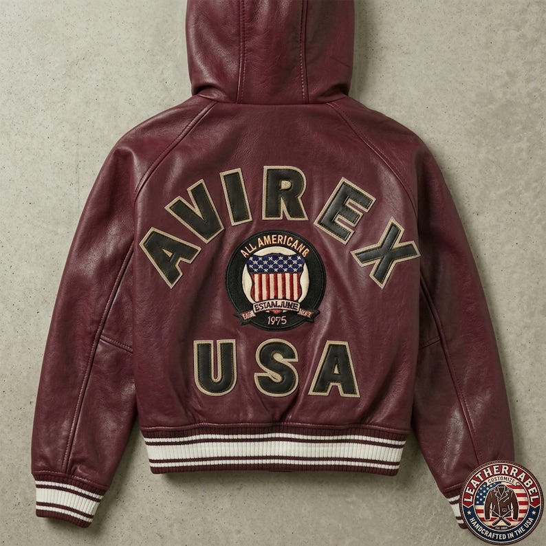 Puede incluir: Una chaqueta de cuero con capucha en color burdeos que presenta el texto AVIREX USA en letras grandes de color negro y crema en la espalda. Un parche circular en el centro muestra un gr&aacute;fico de la bandera estadounidense con el texto ALL AMERICAN ESTABLISHED 1975. La chaqueta tiene pu&ntilde;os y dobladillo acanalados con rayas blancas y burdeos.