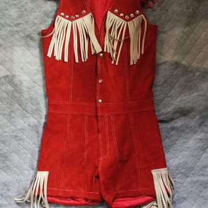 Puede incluir: Un romper rojo brillante sin mangas con detalles de flecos color crema en los hombros y en las aberturas de las piernas. La prenda incluye botones de presi&oacute;n de metal plateado en la parte delantera y un forro interior rojo a juego.