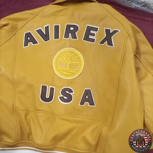 May include: Mustard yellow leather jacket with the word "AVIREX" and "USA" in brown lettering. A circular patch with a tree design is centered between the text. The jacket has a white and yellow striped trim.