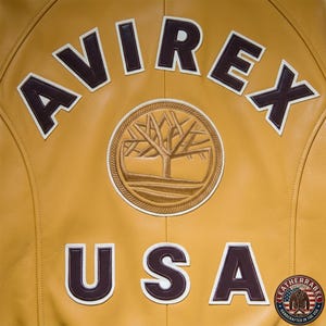 May include: Mustard yellow leather jacket with the word "AVIREX" arched across the top in dark brown letters with white trim. Below is "USA" in similar lettering. A circular emblem with a tree design is centered.