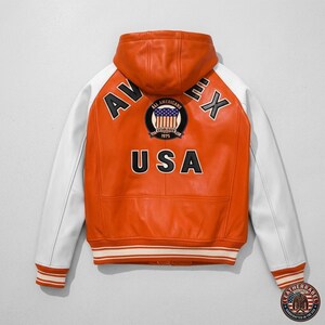 K&ouml;nnte beinhalten: Orange-wei&szlig;e Lederjacke mit Kapuze. Auf der R&uuml;ckseite steht "AV EX USA" und ein kreisf&ouml;rmiger Aufn&auml;her mit der amerikanischen Flagge. Die Jacke hat wei&szlig;e &Auml;rmel und einen gestreiften Saum. Varsity-Stil.