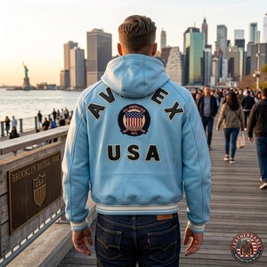 Pu&ograve; includere: Giacca in pelle azzurra con cappuccio, con la scritta "AVIREX" e "USA" sul retro. La giacca ha un bordo a righe bianche e nere in vita e sui polsini. L'immagine &egrave; scattata all'aperto con uno skyline sullo sfondo.