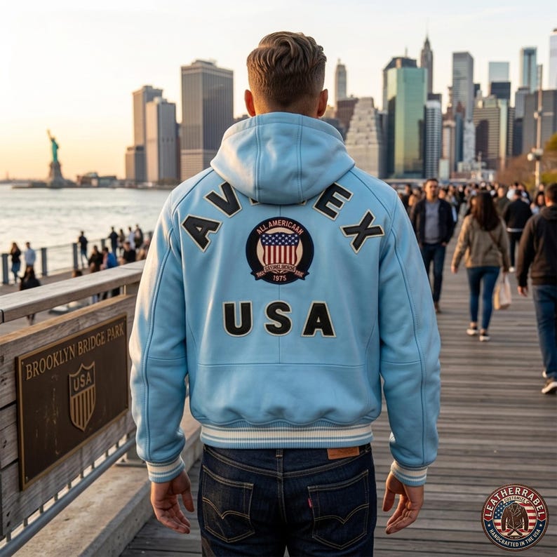Pu&ograve; includere: Giacca in pelle azzurra con cappuccio, con la scritta "AVIREX" e "USA" sul retro. La giacca ha un bordo a righe bianche e nere in vita e sui polsini. L'immagine &egrave; scattata all'aperto con uno skyline sullo sfondo.
