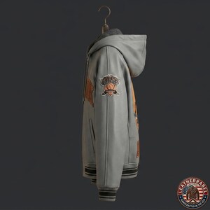 Puede incluir: Una sudadera con capucha de cuero gris claro que presenta capucha, mangas largas con pu&ntilde;os acanalados y un dobladillo con detalles acanalados. La manga muestra un parche bordado de un bal&oacute;n de baloncesto con el texto "KNICKS" y letras naranjas.