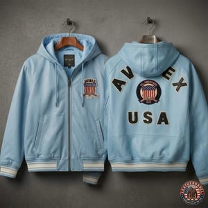 Pu&ograve; includere: Giacca in pelle azzurra con cappuccio e chiusura a zip. Presenta una toppa con un disegno della bandiera americana sul petto e sulla schiena. La giacca ha polsini e orlo a righe bianche e blu. Il testo "AVIREX" e "USA" &egrave; visibile sul retro.