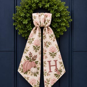 May include: A green boxwood wreath adorned with a long, cream-coloured fabric ribbon featuring a floral pattern of pink flowers and green leaves. The ribbon displays a pink pumpkin, a white pumpkin, and the capital letter H in pink embroidery.