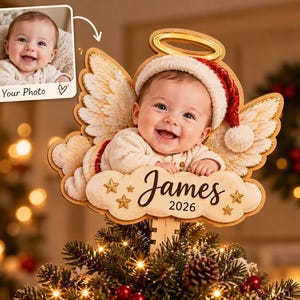May include: A custom Christmas tree topper featuring a photograph of a smiling baby wearing a red and white Santa hat. The baby is framed by wooden angel wings, a gold halo, and a cloud-shaped base with the name James and the year 2026 printed on it.