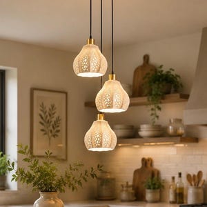 May include: A three-light pendant fixture featuring white, bell-shaped shades with intricate, perforated cut-out patterns. The shades are suspended by black cords from a circular brass ceiling mount, emitting a warm glow from within.