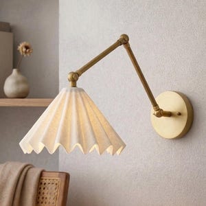 May include: A wall-mounted lamp featuring an adjustable brass arm and a pleated, off-white conical shade. The fixture attaches to the wall with a circular brass plate and extends outward with two hinged joints to position the light source.