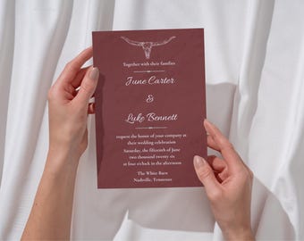 May include: A burgundy wedding invitation with white text, held by hands. The invitation features a steer skull illustration and the names "June Carter & Luke Bennett." Details include the wedding date, time, and location: The White Barn, Nashville, Tennessee.