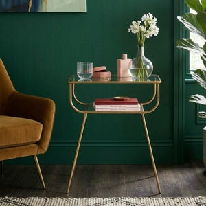 Op de afbeelding: Een goudkleurige bijzettafel met een glazen blad en een onderste plank. De tafel bevat een helder glas, een roze doos, een roze parfumfles, een vaas met witte bloemen en twee boeken. De tafel heeft een gebogen ontwerp en staat tegen een donkergroene muur.