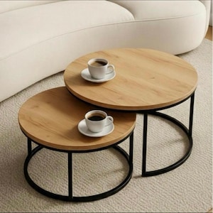 May include: Two round nesting coffee tables with light wood grain tops and black metal frames. Each table has a cup of coffee on a white saucer. The tables are on a light-colored rug.