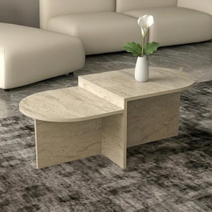 Peut inclure: Une table basse beige moderne avec un design unique à deux niveaux. La table présente un bord arrondi d'un côté et une forme rectangulaire de l'autre. Un vase blanc avec une fleur blanche et des feuilles vertes est posé dessus.