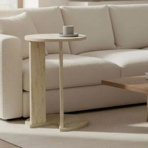 Puede incluir: Una mesa auxiliar redonda de color claro con una sola pata y una pequeña taza y platillo grises encima. La mesa es de madera clara y está colocada junto a un sofá color crema. La mesa mide aproximadamente 61 cm de altura.