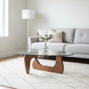 May include: A living room with a glass-topped coffee table with a distinctive wooden base. The table sits on a white rug with a geometric design. A grey sofa with beige cushions and a white floor lamp are also visible in the room.