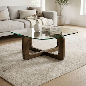 Puede incluir: Una mesa de centro cuadrada con una tapa de vidrio transparente y una base de madera marrón oscuro. La base tiene un diseño en forma de X. La mesa está en un salón con un sofá gris y una alfombra beige.