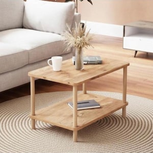 Puede incluir: Mesa de centro de madera clara de dos niveles. La mesa tiene una parte superior rectangular y un estante inferior, sostenidos por cuatro patas cilíndricas. Una taza blanca, un jarrón con flores secas y un libro están encima.
