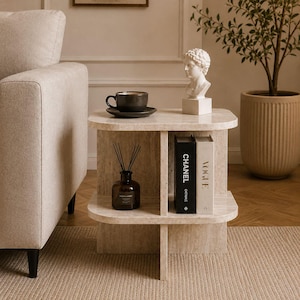 May include: A two-tiered, beige stone side table with rounded edges. A black coffee cup and saucer sit on top, along with a white bust and two books. A dark brown diffuser bottle and a potted plant are also visible.