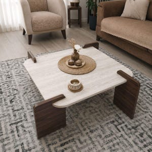 Pode incluir: Uma mesa de centro quadrada moderna com um tampo de pedra clara e pernas de madeira escura. A mesa está numa sala de estar, com um tapete, um sofá e uma poltrona visíveis. Há objetos decorativos na mesa.