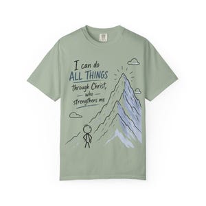 May include: A sage green short-sleeve t-shirt featuring a graphic of a stick figure standing before a mountain. The text reads: I can do ALL THINGS through Christ, who strengthens me. The mountain is rendered in shades of blue and white with clouds.
