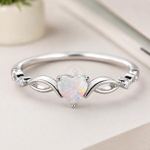 May include: A silver ring featuring a heart-shaped opal gemstone. The opal has a white base with iridescent flashes of color. The band has a delicate design with small, clear gemstones and decorative swirls.