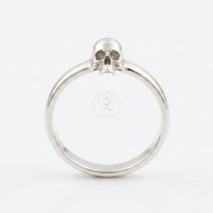 May include: A silver ring featuring a skull design. The skull is positioned at the top of the ring, with a detailed carving of the skull's features. The ring band is smooth and rounded, creating a minimalist aesthetic.