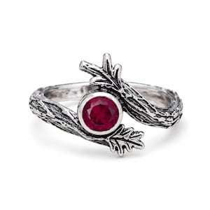 Pode incluir: Um anel de prata com uma banda texturizada que imita ramos de árvore com folhas nas extremidades. O centro do anel apresenta uma pedra preciosa redonda, facetada e de cor vermelho-escuro, engastada num bisel de prata liso, estilo bypass.