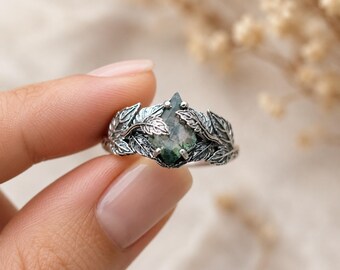 May include: A silver ring featuring a teardrop-shaped, moss-green gemstone. The ring is adorned with detailed leaf designs that wrap around the stone. The ring's intricate design and natural elements create a unique and elegant piece of jewelry.