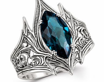 May include: A silver ring featuring a large, marquise-cut dark blue gemstone set in the center. The band is crafted with intricate, raised scrollwork and filigree patterns that extend upward into pointed, crown-like prongs around the stone.