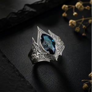 May include: A silver-toned ring with a large, oval, blue gemstone. The ring has an ornate, gothic-inspired design with intricate detailing and pointed edges. The gemstone appears to have an etched design. The ring is displayed on a black surface.