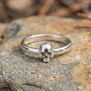 May include: A silver ring with a skull motif. The skull is the central design element, with clear eye sockets, nose, and jaw details. The ring's band is polished, offering a gothic or alternative aesthetic.