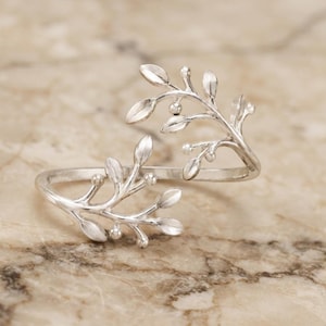 May include: A silver-toned open ring featuring two mirrored branches with small, smooth leaves and tiny spherical buds. The band is crafted from a continuous metal wire that splits into two delicate, organic-shaped sprigs that meet at the centre.