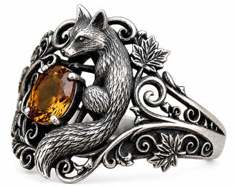 May include: A silver ring featuring a sculpted fox figure and an oval-cut amber-colored gemstone. The band is decorated with intricate metal scrollwork and detailed maple leaf motifs, creating a textured, oxidized finish throughout the piece.