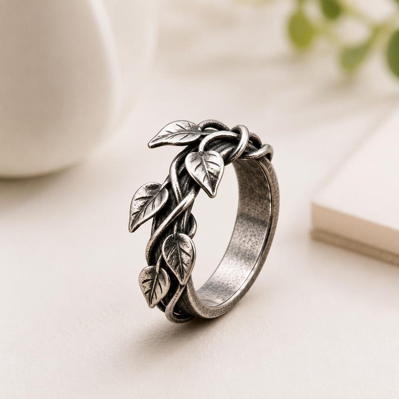 May include: A silver-toned metal ring featuring a band designed to resemble intertwined vines with small, raised leaf details. The metal has a darkened, oxidized finish in the recessed areas to highlight the texture of the leaves and stems.