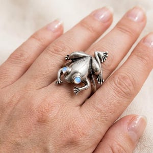 May include: A silver-toned metal ring shaped like a frog, featuring two small, round, iridescent moonstone-like cabochons as eyes. The frog design shows detailed limbs and a smooth, polished back, with the band extending from the underside.